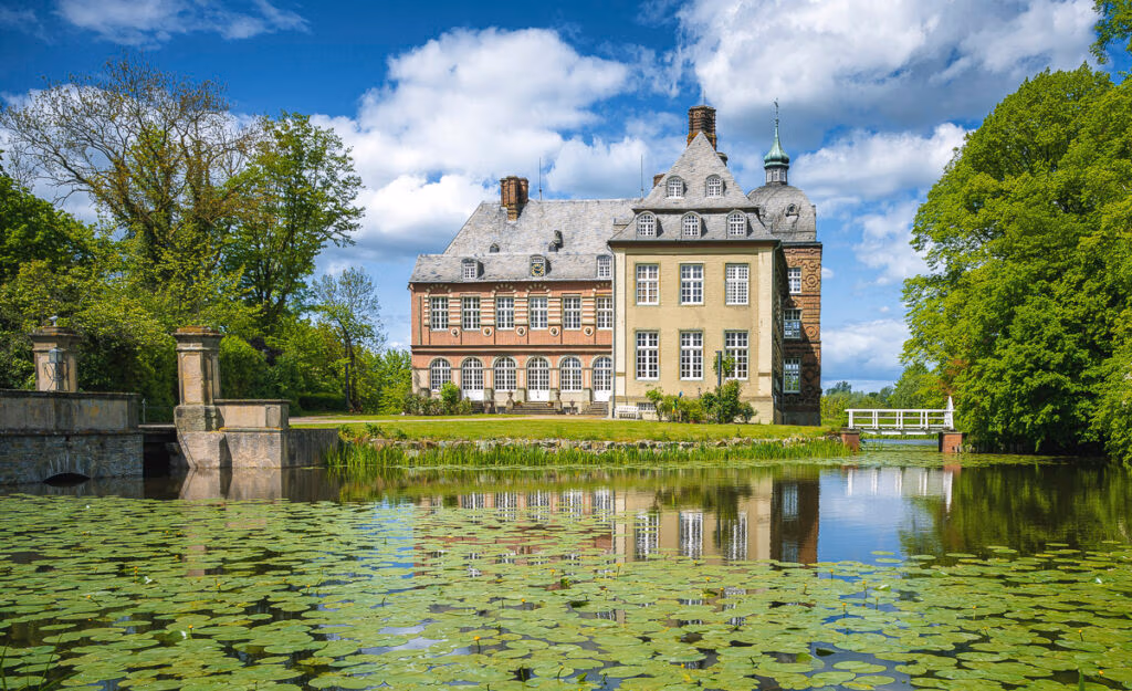 Schloss Hovestadt: Historisches Herrenhaus mit Teich, Seerosen und Bäumen unter blauem Himmel.