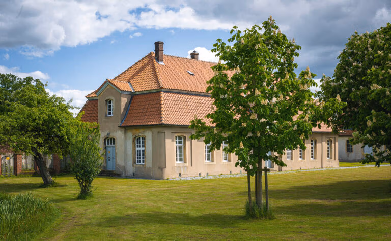 Ein beiges Haus mit rotem Ziegeldach, wie Schloss Hovestadt, steht inmitten grüner Bäume.