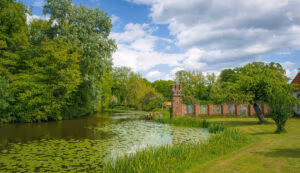 Ein Teich mit Seerosen neben der Backsteinmauer von Schloss Hovestadt, inmitten üppiger grüner Bäume.