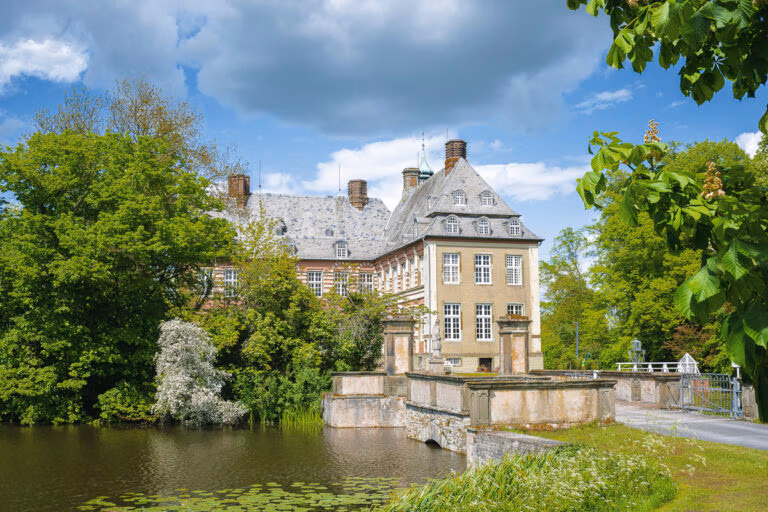 Historisches Herrenhaus aus Stein, Schloss Hovestadt, mit steilen D&auml;chern, B&auml;umen und einem Teich unter blauem Himmel.