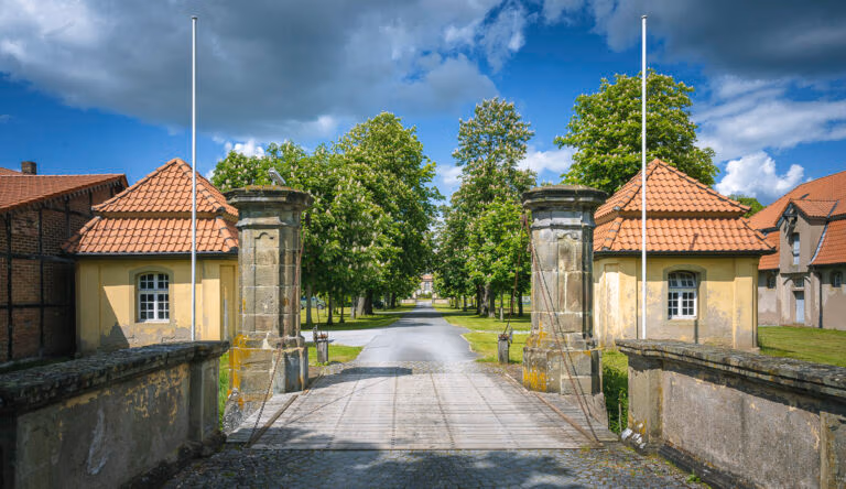 Das Steintor von Schloss Hovestadt mit seinen roten Ziegeld&auml;chern &ouml;ffnet sich zu einer von B&auml;umen ges&auml;umten Stra&szlig;e und blauem Himmel.