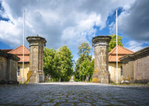 Steinernes Eingangstor zum Schloss Hovestadt mit Säulen, Kopfsteinpflasterweg und roten Ziegeldächern.