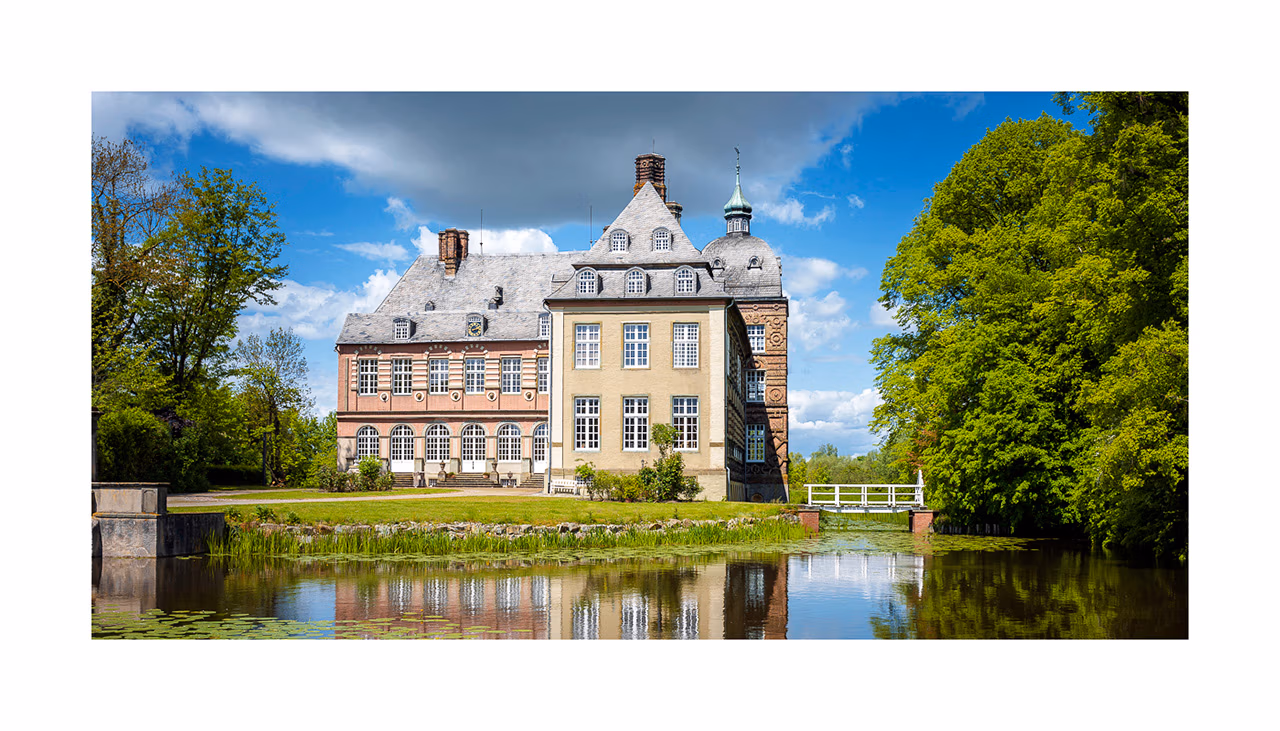Historisches Anwesen Schloss Hovestadt mit einem Teich, eingebettet zwischen Bäumen unter einem teilweise bewölkten Himmel.