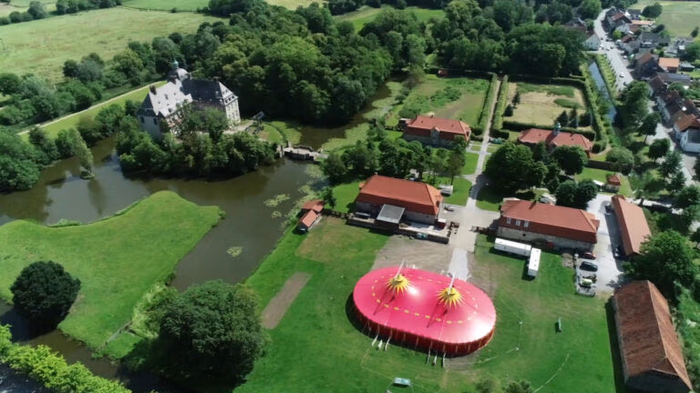 Luftaufnahme eines roten Zirkuszeltes in der Nähe von Schloss Hovestadt, umgeben von Bäumen und Wasser.